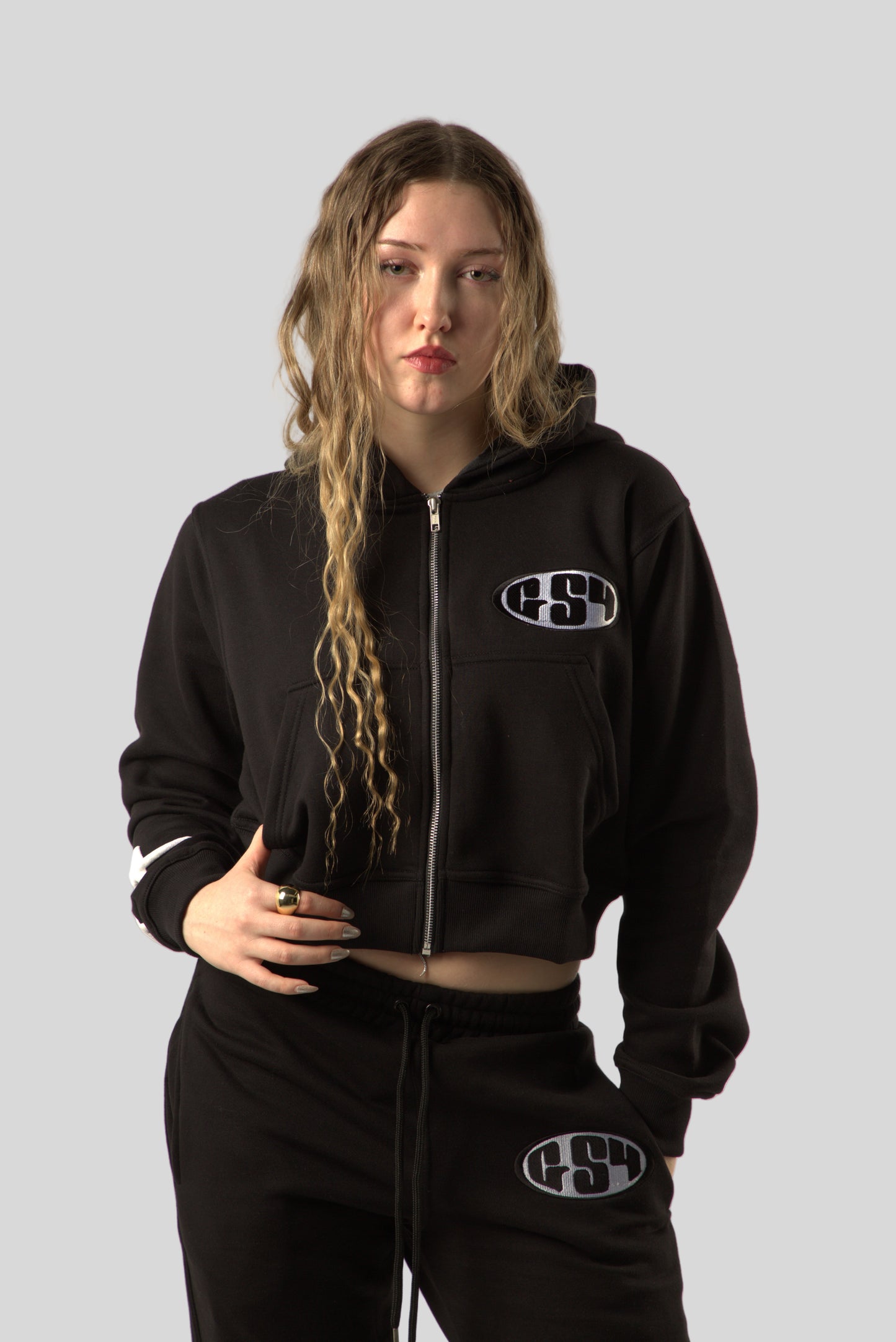 A woman wearing a black tracksuit with a bold white puff print on the jumper. The tracksuit includes a hood with the same print.